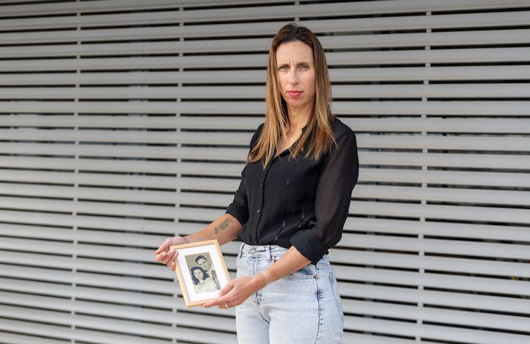 A woman in a black shirt and jeans holds out a framed photograph.